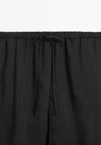 Black elastic waistband with a tied drawstring on lightweight, textured fabric, part of loose-fitting pants or shorts.