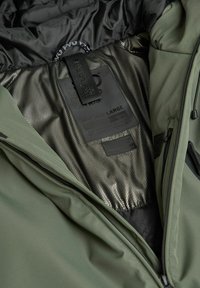 Wnętrze oliwkowej kurtki z czarną podszewką, kieszenią siatkową, metką rozmiaru "LARGE" oraz szczegółami z teksturowanej tkaniny.