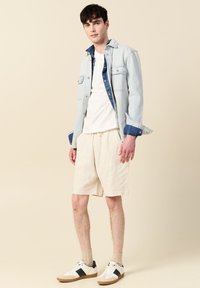 Vaaleansininen farkkutakki sinisillä yksityiskohdilla, valkoinen T-paita, beigekoriset shortsit ja valkoiset lenkkarit mustilla yksityiskohdilla. Yksinkertainen, rento yhdistelmä.