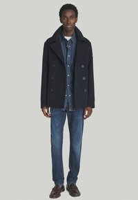 Cappotto peacoat in lana blu scuro con grandi bottoni, indossato sopra una camicia di denim, abbinato a jeans sbiaditi e scarpe in pelle scura.