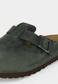 Σκούρο πράσινο δερμάτινο τσόκαρο με ρυθμιζόμενο λουράκι και μεταλλική πόρπη, με περιμετρική σόλα και σουέντ φόδρα.