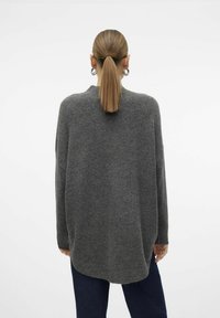 Szary, oversize'owy sweter z dzianiny z okrągłym dekoltem, długimi rękawami i zaokrąglonym dołem, zestawiony z ciemnoniebieskimi spodniami dżinsowymi.