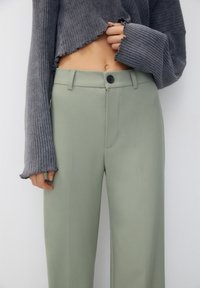 Pull en maille côtelée gris, cropped, avec des bords effilochés, associé à un pantalon taille haute vert clair, ajusté, doté d'une fermeture à bouton et de poches latérales.