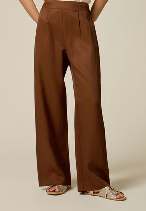 Pantalones marrones de talle alto y pierna ancha combinados con sandalias planas beige tejidas sobre un fondo claro y liso.