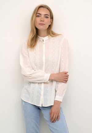 Weiße, semi-transparente Bluse mit gestickten Blumenmustern, Stehkragen, Knopfverschluss und langen Ärmeln. Getragen mit blauen Jeans.