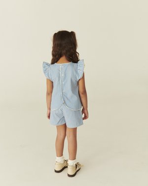 Jeune fille aux cheveux longs, vêtue d'une robe à carreaux bleus et blancs et de chaussures blanches, vue de dos sur un fond uni.