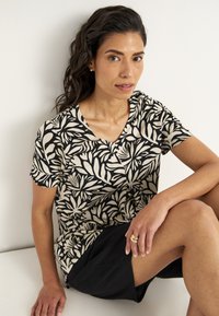 Korte mouw top in crème met zwart bladerenpatroon; v-halslijn; model zit in zwarte shorts, met een gouden ring om de rechterhand.