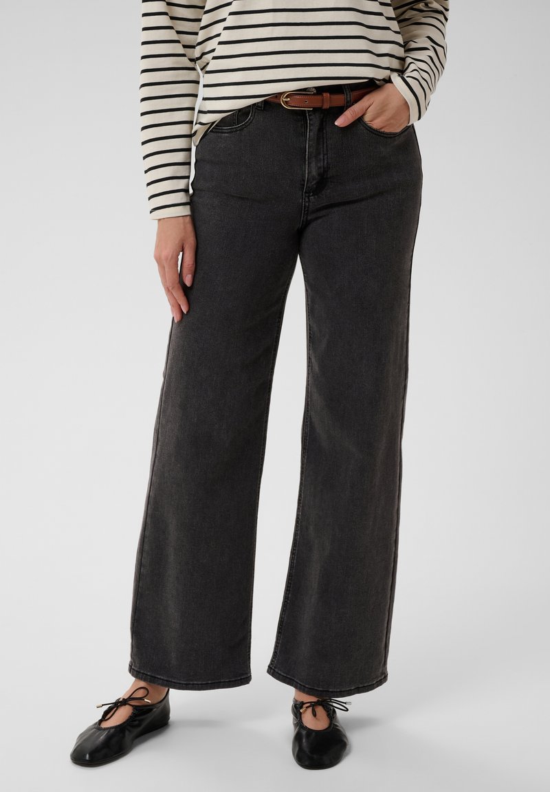 Personne portant un jean noir à jambes larges, une ceinture marron, des ballerines noires et une chemise beige rayée à manches longues, une main dans la poche.