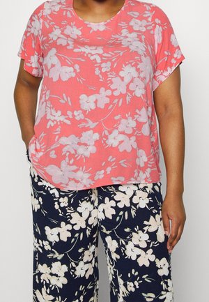 Persoon draagt een koraalkleurige korte mouw top met lichtroze bloemenprint en marineblauwe wijde broek met witte bloemmotieven.