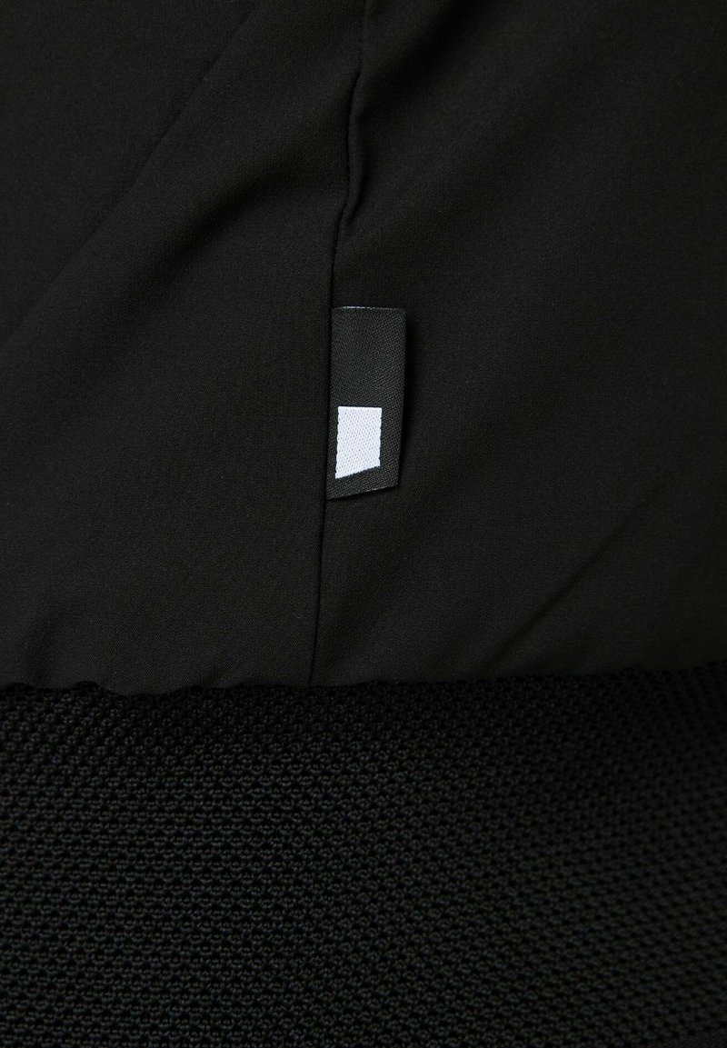 Czarny materiał o teksturowanej matowej powierzchni, z małą białą metką i gładkimi sekcjami. Kontrast między górną gładką częścią a dolną teksturowaną.