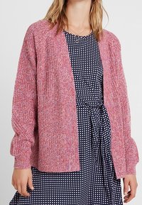 Cardigan rosa a trama con una vestibilità ampia, apertura frontale e maniche lunghe, abbinato a un vestito blu navy a pois. Materiale in maglia morbida.