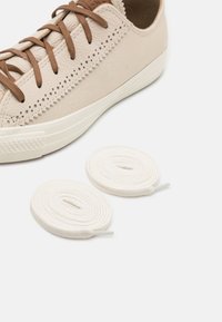 Beige színű sneaker, amely texturált vászonból és sima bőrből készült kiegészítőkkel rendelkezik, mellette két fehér pamut cipőfűző, szépen feltekerve.