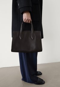 Donkerbruine leren totebag met verlengde handvatten, gladde textuur, rechthoekige vorm, vastgehouden door een persoon in donkere kleding.