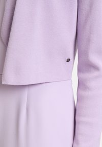 Hellpurpurner, gestrickter Cardigan mit weicher Textur, offenem Frontdesign und einem kleinen schwarzen Logoakzent am Ärmel.