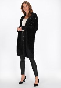 Donna che indossa un lungo cardigan nero peloso con dettagli in perle, un top bianco con pizzo, pantaloni in pelle neri e tacchi alti neri, in piedi.