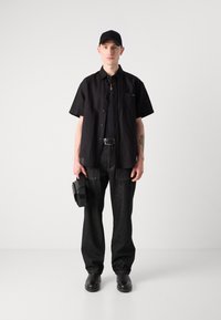 Camisa negra de manga corta, camiseta negra debajo, pantalones vaqueros oscuros, gorra negra, sosteniendo un bolso circular negro, zapatos de cuero.