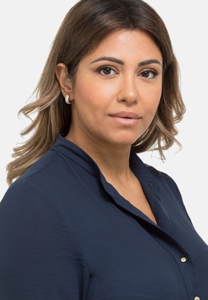 Camicia blu navy con una texture morbida e colletto, dotata di bottoni metallici e una vestibilità comoda, indossata con un piccolo orecchino texturizzato.