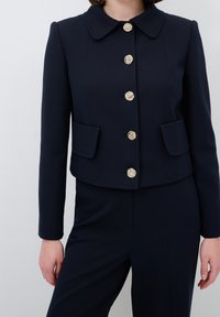Marinblå kort jacka med rund krage, guldknappdetaljer och två framfickor. Slät textur, skräddarsydd passform.