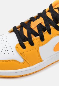 Vit och orange sneaker med perforerat tåparti, svarta snörningar och slätt syntetiskt material. Har en vadderad plös och orange detaljer.