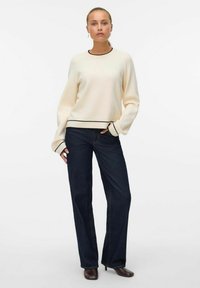 Krēmkrāsas džemperis ar melnu apdari, garām piedurknēm un brīva piegriezuma, kopā ar tumši zilām taisnām džinsa biksēm un kurpēm ar papēžiem.