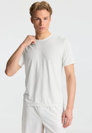 Ung mand med blondt hår iført en enkel hvid kortærmet t-shirt og hvide bukser, stående mod en lys baggrund.