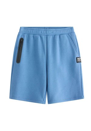 Teenage dreng står iført en mørk hættetrøje, blå shorts, hvide sneakers, stribede sokker og en blå mønstret baseballkasket mod en ensfarvet baggrund.