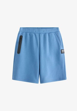 Teenage dreng står iført en mørk hættetrøje, blå shorts, hvide sneakers, stribede sokker og en blå mønstret baseballkasket mod en ensfarvet baggrund.