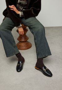 Zwarte leren instapschoenen met een gouden kettingaccent, gedragen met donkerbruine sokken, gecombineerd met wijde denim jeans en een geruit overhemd.