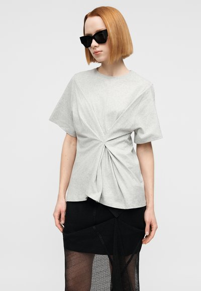 Γκρι ανοιχτόχρωμο κοντομάνικο μπλουζάκι με σχέδιο στριμμένο μπροστά. Φορεμένο με μια μαύρη φούστα σε στρώσεις που διαθέτει διακοσμητικά από δίχτυ και ύφασμα. Μαύρα γυαλιά ηλίου.