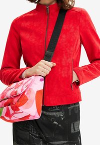 Giacca in ecopelle rossa con collo alto e chiusura con zip, abbinata a una borsa rosa con motivo floreale e tracolla nera.