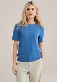 Zils īsām piedurknēm svīteris ar teksturētu rakstu, ribotas apkakles un apakšmalas, kopā ar gaiši bežām biksēm. Gluda, viegla auduma.