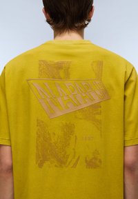 Sennepsgul t-shirt med et stort, tekstureret "NAPAPIJRI" logo i brun på bagsiden, med "1987" trykt let under.
