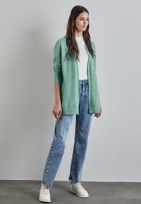 Hellgrüne Strickjacke mit gerippten Bündchen, kombiniert mit hellblauen geraden Jeans und weißen Sneakers auf einem strukturierten grauen Boden.