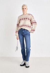 Beige strikkegenser med maroon geometrisk mønster, avslappet passform. Kombinert med blå jeans og sølvfargede snøresko, holder en metallisk veske.