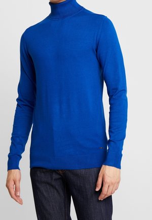 Man met een felblauwe coltrui gecombineerd met donkerblauwe spijkerbroek, staand tegen een eenvoudige lichte achtergrond.