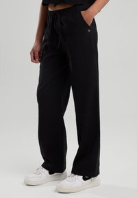 Mustat leveät housut kevyestä kankaasta, joissa on naruvyötärö, sivutaskut ja teksturoitu pinta. Yhdistetty valkoisiin lenkkareihin.