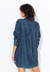 Robe chemise en denim bleu foncé avec manches longues, poignets boutonnés et détail de plis au dos. Sa texture douce et son design décontracté renforcent sa polyvalence.