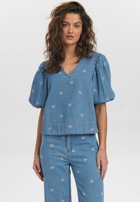 Ljusblå denimtop med puffärmar, V-ringning, blommig brodyr och en kortad fåll. Matchas med ljusblå denimbyxor.