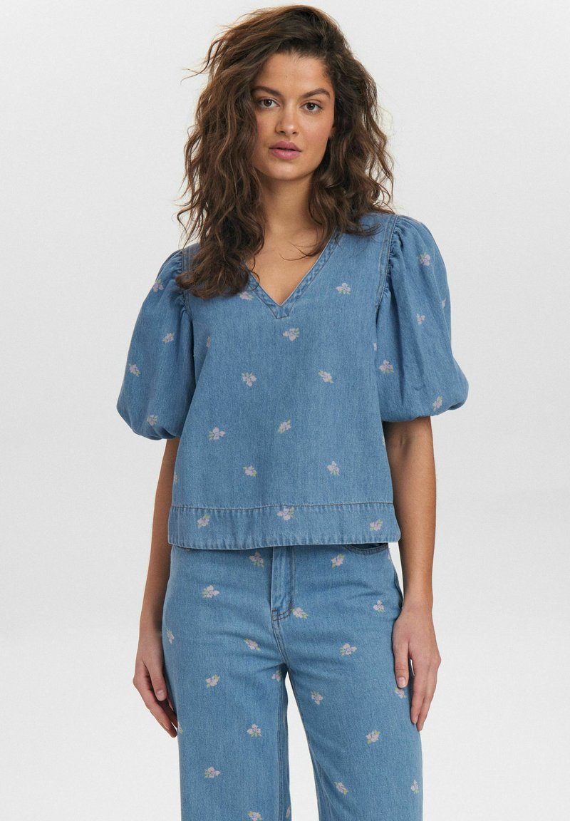 Ljusblå denimtop med puffärmar, V-ringning, blommig brodyr och en kortad fåll. Matchas med ljusblå denimbyxor.