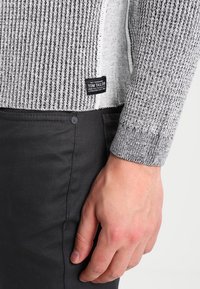 Pull rayé gris et noir avec une texture côtelée et une étiquette portant la mention "Tom Tailor". Porté avec un pantalon noir, montrant les détails des poignets.