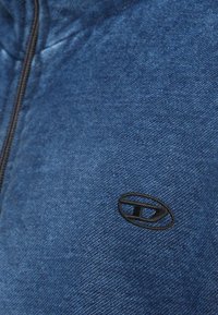 Modra, teksturirana jopa z zadrgo in črnim logotipom na levi strani. Tkanina ima mehak, tkan videz in gladko zadrgo.