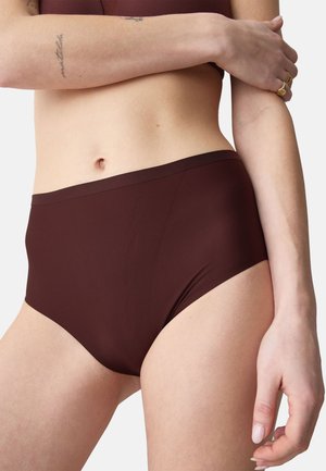 Vrouw die donkerbruin high-waist ondergoed en bijpassende bh draagt, met getatoeëerde armen gekruist over haar torso tegen een effen achtergrond.