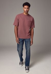 T-shirt a maniche corte, color malva, con taschino sul petto, abbinato a jeans blu e sneakers nere, creando un look informale.
