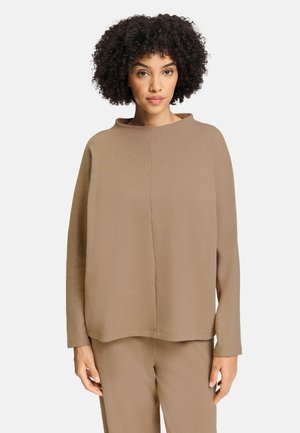 Kvinde med krøllet hår iført en løstsiddende beige langærmet top og matchende bukser, stående mod en ensfarvet hvid baggrund.