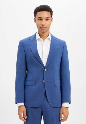 Jonge man met kort krullend haar die een aansluitend blauw pak en een wit overhemd draagt, staand tegen een effen witte achtergrond.