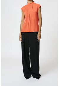 Blouse sans manches corail avec col mandarin, ornée de plis à l'avant. Associée à un pantalon noir large, conçu dans un tissu lisse.