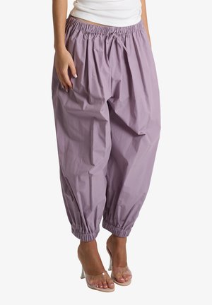 Donna con pantaloni larghi color lilla con vita e polsini elastici abbinati a sandali trasparenti con tacco alto, in piedi su sfondo bianco.