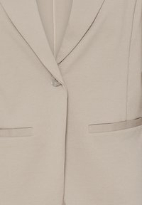 Blazer beige clair avec revers crantés, fermeture par un bouton unique et deux poches horizontales fendues sur le devant.