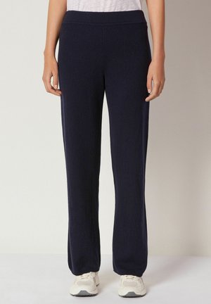 Femme portant un pantalon droit bleu marine et des baskets blanches, se tenant devant un fond clair uni.