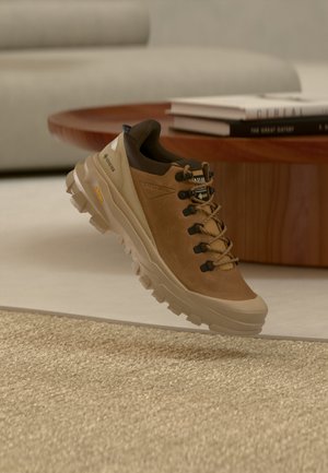 Καφέ ορειβατικό παπούτσι με παχιά σόλα Vibram και ετικέτα Gore-Tex, τοποθετημένο πάνω από μπεζ χαλί κοντά σε ένα ξύλινο τραπέζι με στοιβαγμένα βιβλία.